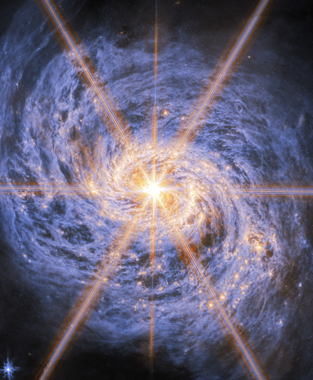 A spiral galaxy shown in mid-infrared light. The image is dominated by an extremely bright glow from the galaxy’s nucleus. Six large and two smaller rays of light emit from the center, which are diffraction spikes created by the telescope’s optics. The galaxy’s spiral arms are visible by two lines of glowing orange bubbles which whirl out into the disc. Swirling blue clouds of dust make up the rest of the galaxy.