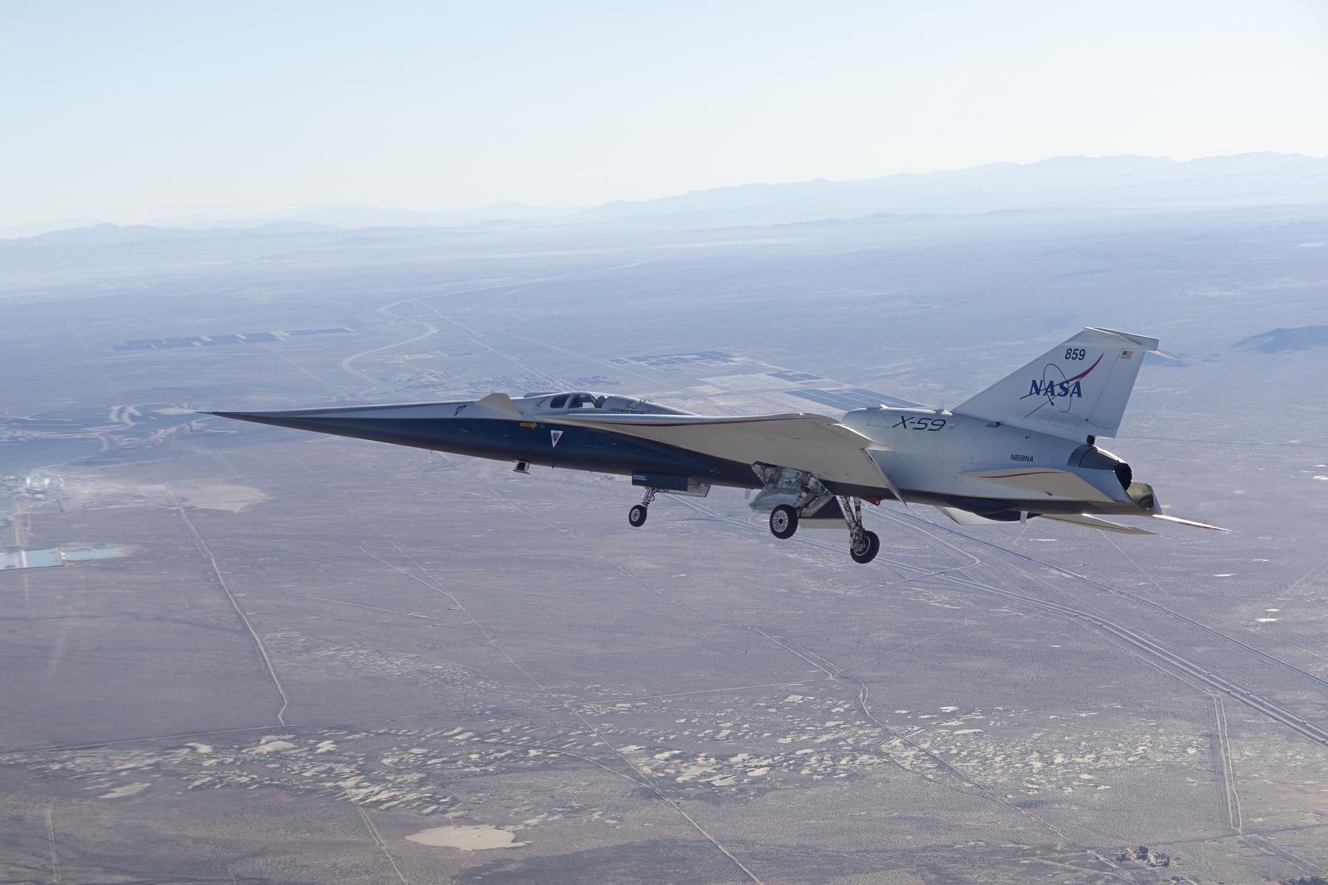 NASA’s X-59 is seen in flight, with a blue sky and mountains behind it and land below it. The aircraft’s long nose and distinct silhouette are visible as it ferries to NASA’s Armstrong Flight Research Center in Edwards, California