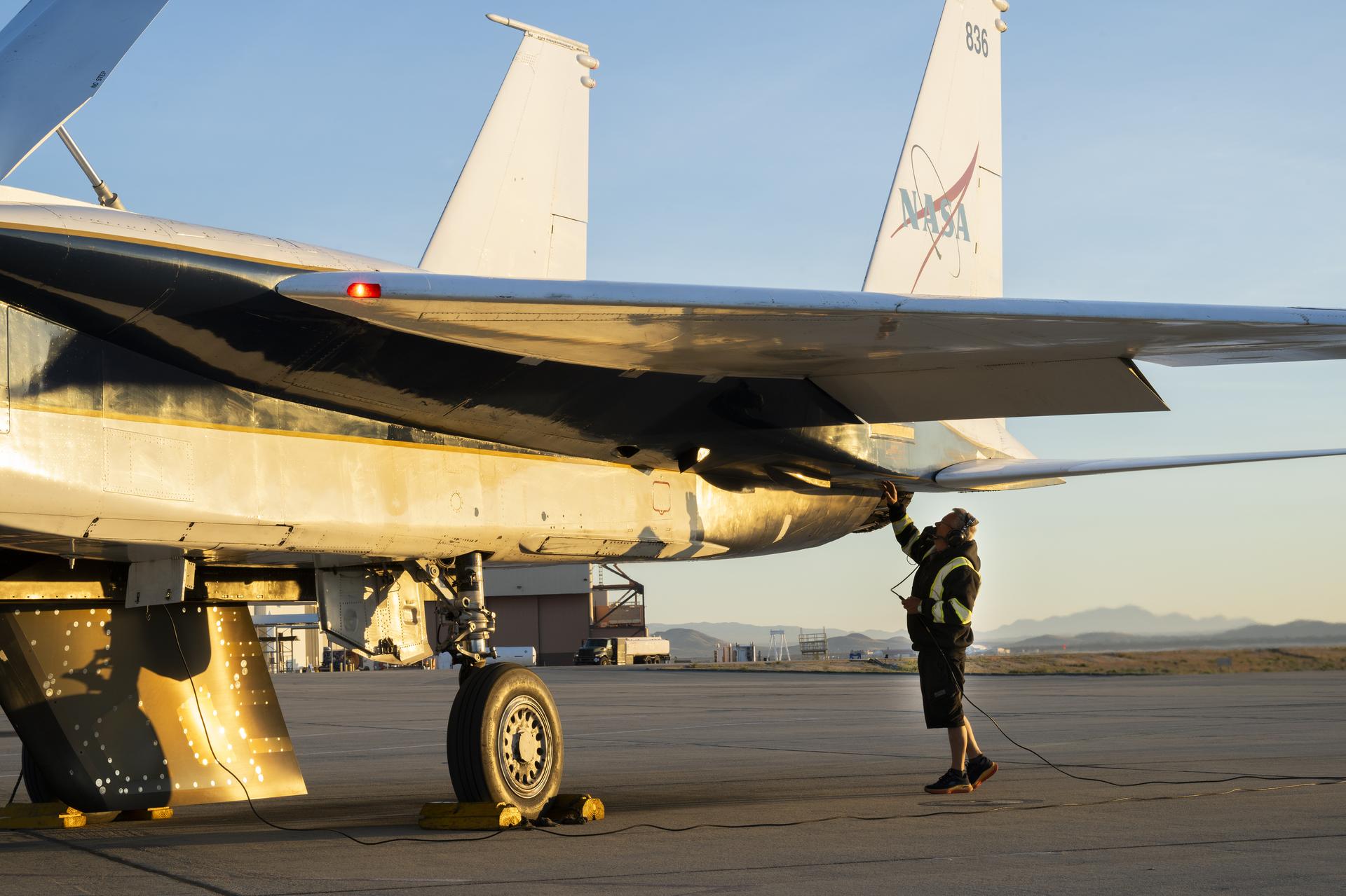 The back of a white jet aircraft’s wing and tails are seen in this image. One wheel and an attached test object, shaped like a wing, hangs from the bottom of the jet. A man in a black jacket, with yellow reflective coating, stands underneath and raises one hand to the aircraft. The man is wearing ear protection. Desert sand and mountains are seen in the background.