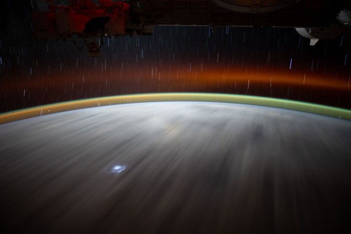 A time-lapse image of Earth from space shows two layers of airglow above Earth's surface: a band of green near the surface and a band of red higher up, with a gap of dark space in between. Star trails are visible in the background above Earth. On Earth, clouds appear as white streaks, with a flash of lightning near the bottom.