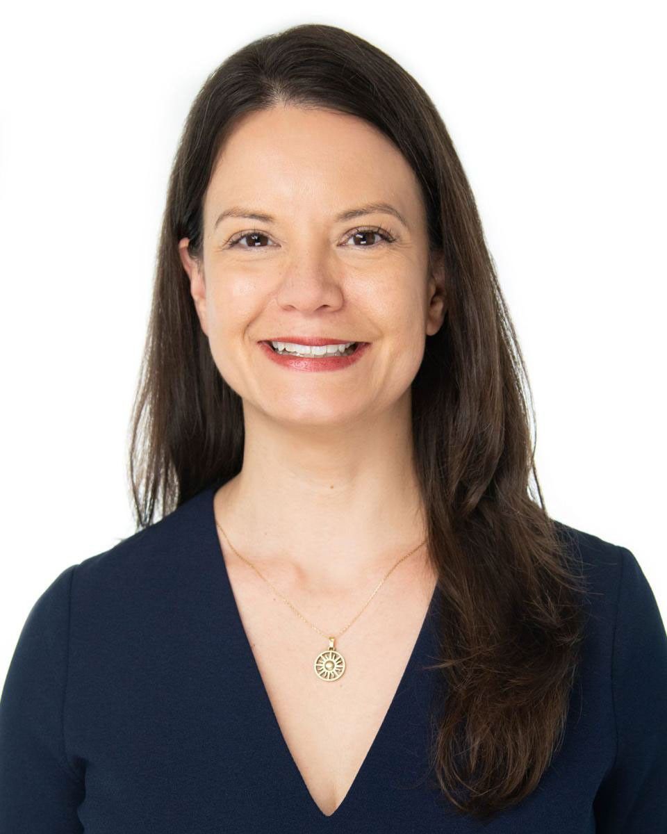 Photo of a smiling woman with brown hair, a navy blue blouse, and pendant necklace.