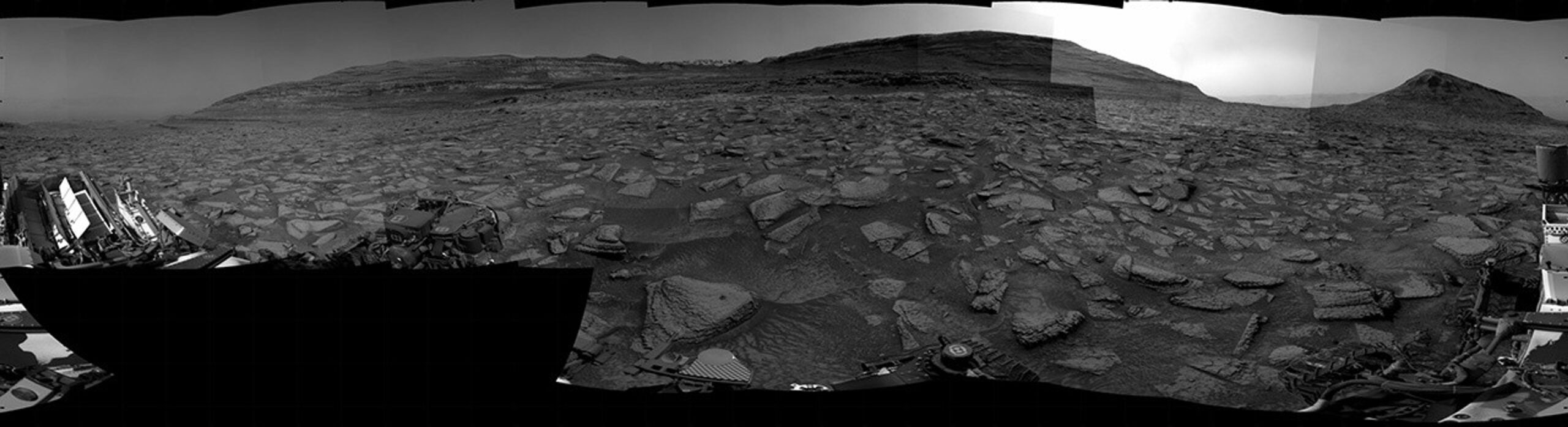 A grayscale panorama of the Martian surface, possibly at night or in twilight, shows a wide field of flat, dark gray terrain dotted with multiple jagged, medium-sized rocks stretching into the distance, where hills rise from the ground on the horizon. At center-right, a bright light behind a hill illuminates part of the sky, while the rest of the scene is fairly dark. Portions of the Curiosity rover are visible along the bottom of the image, including wheels in the lower-right corner.