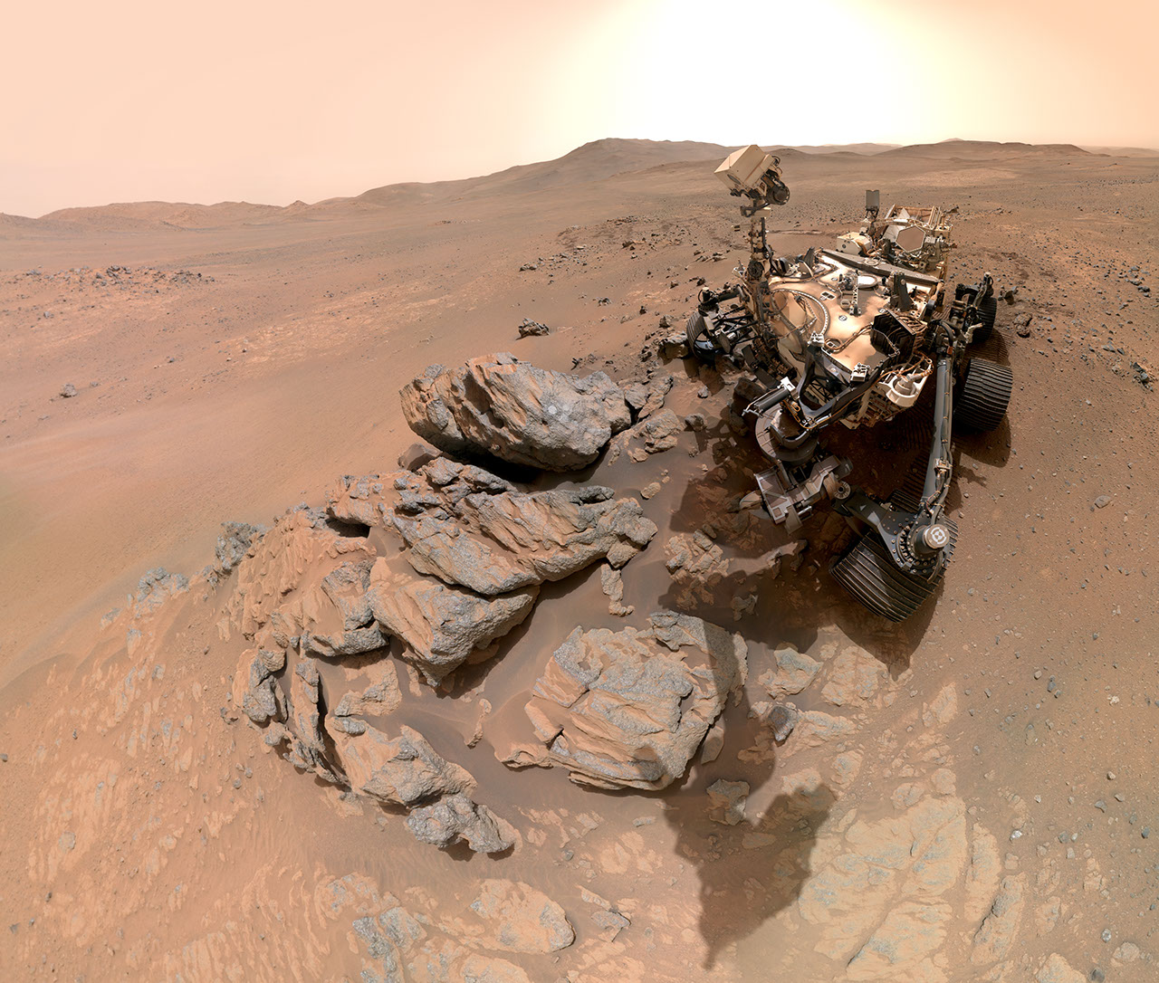 The head of a robotic rover looks down at a rocky outcrop in the foreground. The dusty, orange-red Martian surface stretches away toward the crater rim in the distance. The Sun seems to bloom in a hazy sky.