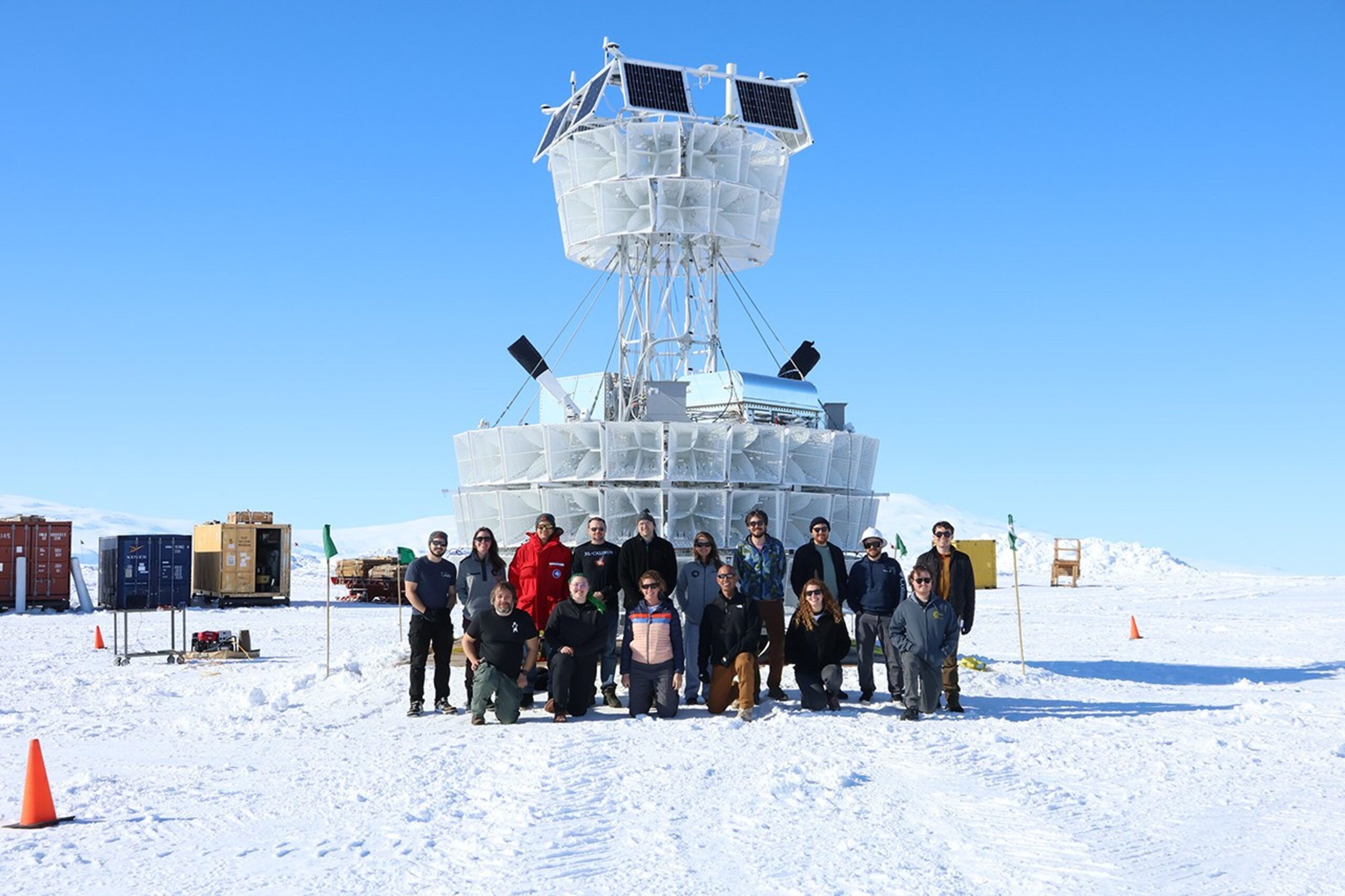Two rows of people in front of a tall structure consisting of rows of white components placed in a circle, topped by metallic containers and an additional circular layer of white structures supported by a tower.