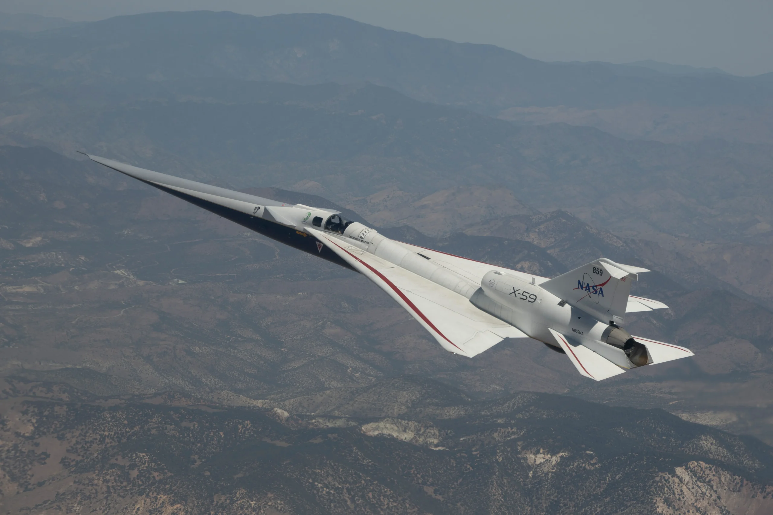 NASA’s X-59 quiet supersonic research aircraft flying above mountain ridges in California during a test flight on May 12, 2026. The aircraft is viewed from above, showing its elongated nose, swept wings, and white fuselage against layered mountains and valleys below.