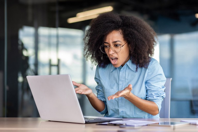 An aghast woman gestures in frustration at a laptop computer on the desk in front of her.