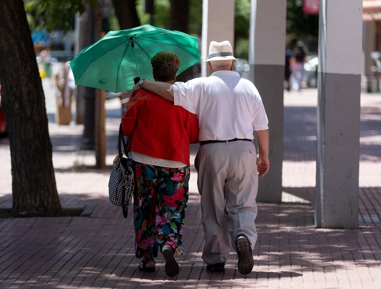 An elderly man with his arm around the shoulders of an elderly woman holding an umbrella walk together down a sunny street.