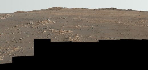 Undulating terrain of reddish-brown soil and loose rocks stretches toward a rugged horizon. The sky above is hazy and pale. The image’s bottom edge is a jagged black border, indicating the edges of the frames stitched together to create the mosaic.