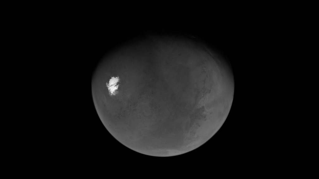 A grayscale view of the Martian globe against a black background, showcasing a brilliant white polar ice cap in sharp contrast with the planet's textured, cratered terrain.