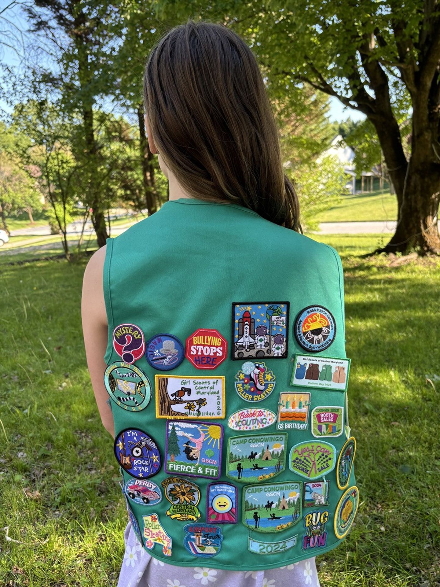 A young Girl Scout stands facing away from the camera, wearing her Junior Girl Scouts vest. The back of the vest is filled with a number of colorful and intricately embroidered patches. The upper right is the PUNCH Ancient and Modern Sun Watching patch earned at this event