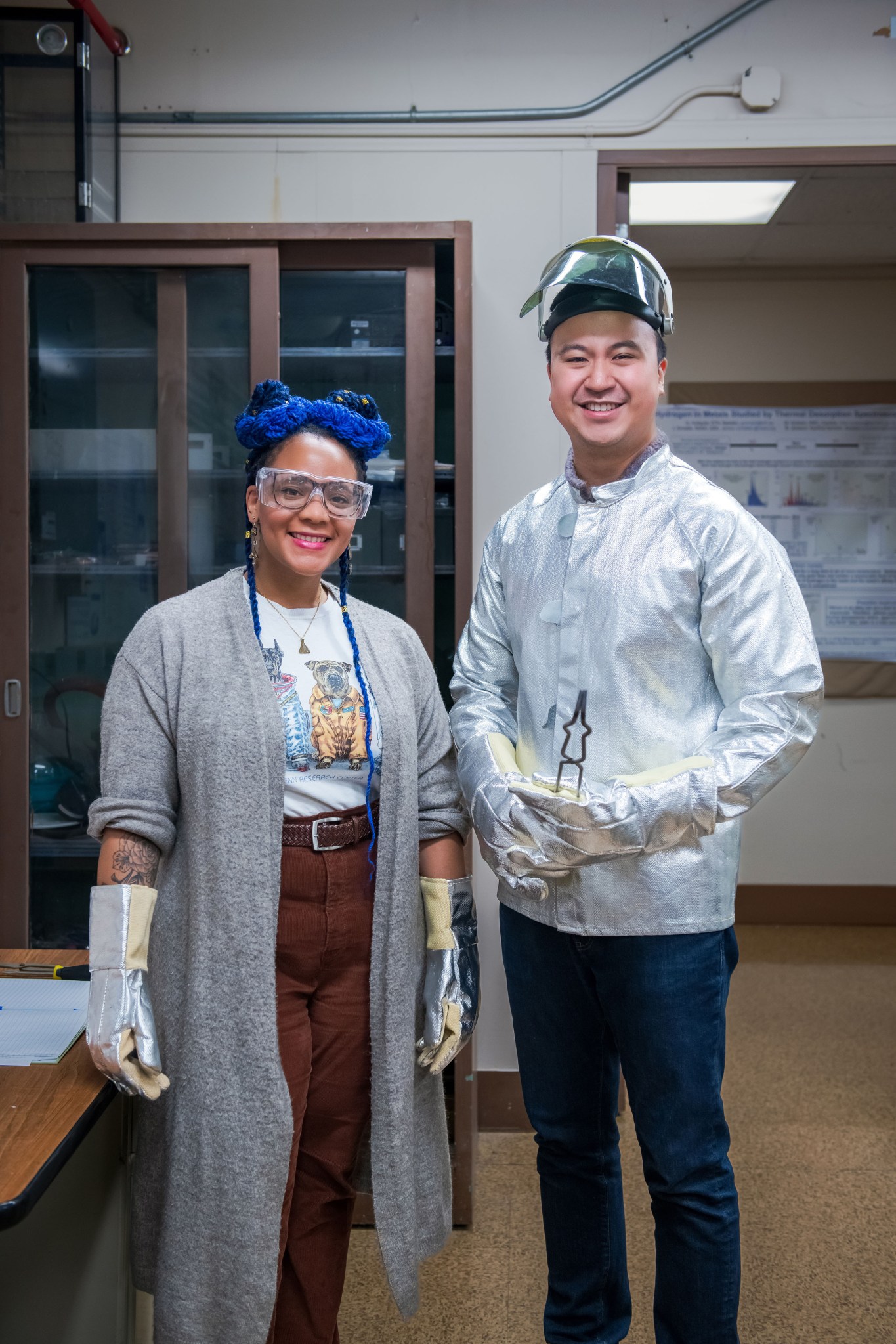 Two researchers pose for a portrait inside a laboratory at NASA Glenn. The researcher on the left is wearing a long grey sweater, silver protective gloves, and safety glasses. The researcher on the right is wearing a silver protective coat, helmet, and gloves and is holding large tongs.