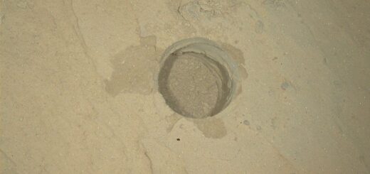 A close-up view of a perfectly circular drill hole in light tan Martian bedrock, created by the Curiosity rover. The shallow hole is filled with loose, pale, powdery rock cuttings. The surrounding rock surface is mostly flat and dusty, featuring subtle ridges, faint cracks, and a few small, scattered pebbles.