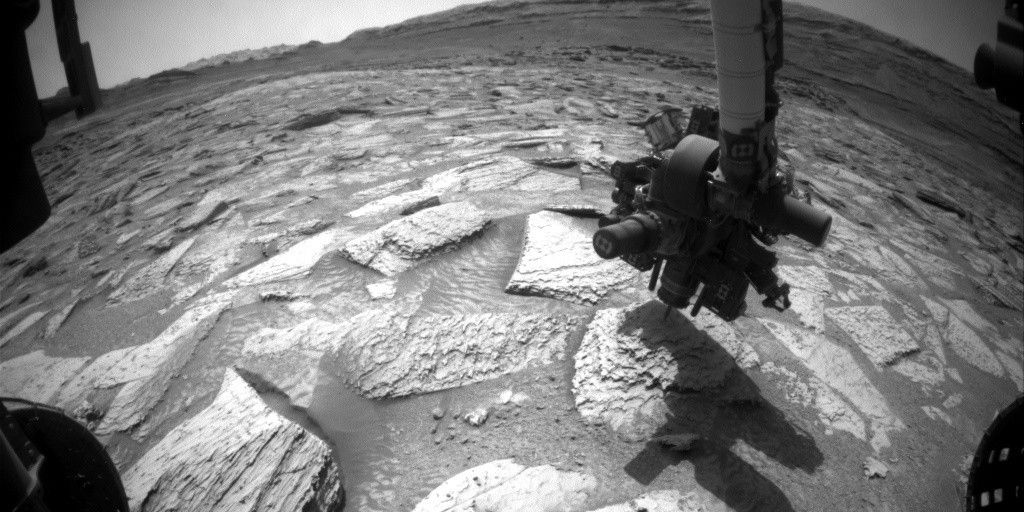 A black and white photo taken from the perspective of the Curiosity rover on Mars. In the center, the rover's robotic arm extends downward, hovering its instrument turret over a flat, light-colored rock. The arm casts a prominent, long shadow across the rocky and sandy terrain in the foreground. Parts of the rover's wheels are visible on the far left and right edges, and a distant, mountainous Martian horizon stretches across the background under a clear sky.