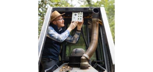 A man in dark gray jeans, a blue checkered long-sleeve, black vest, and straw hat stands on the tread of a black and yellow bulldozer, holding a white sensor box the size of a lunchbox up to the roof in front of the cab. In the background is a green forest.