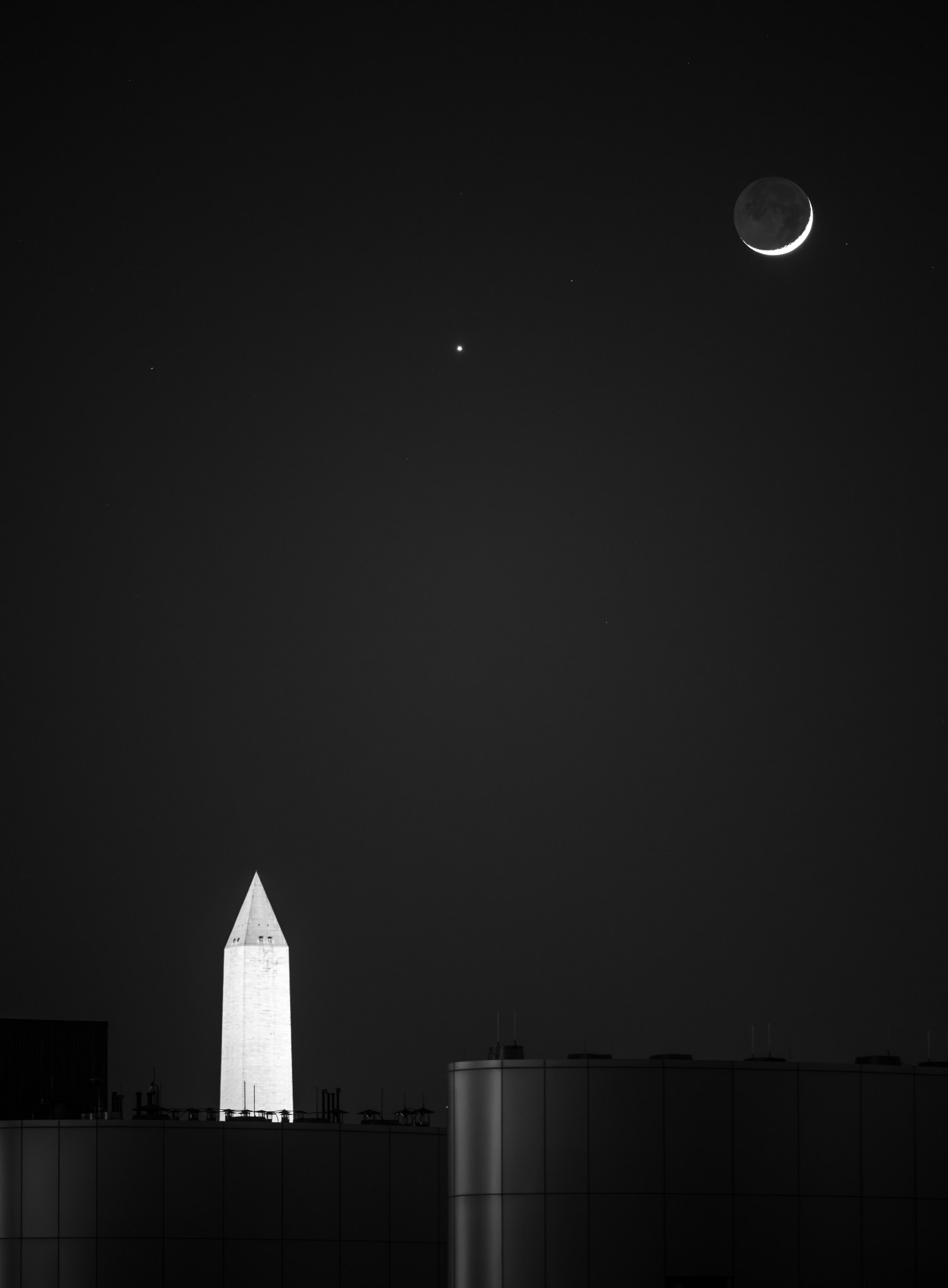 The Moon (right) and Venus (the small dot of light at center) shine above the lit-up Washington Monument at night. Only a small sliver of the Moon is visible.
