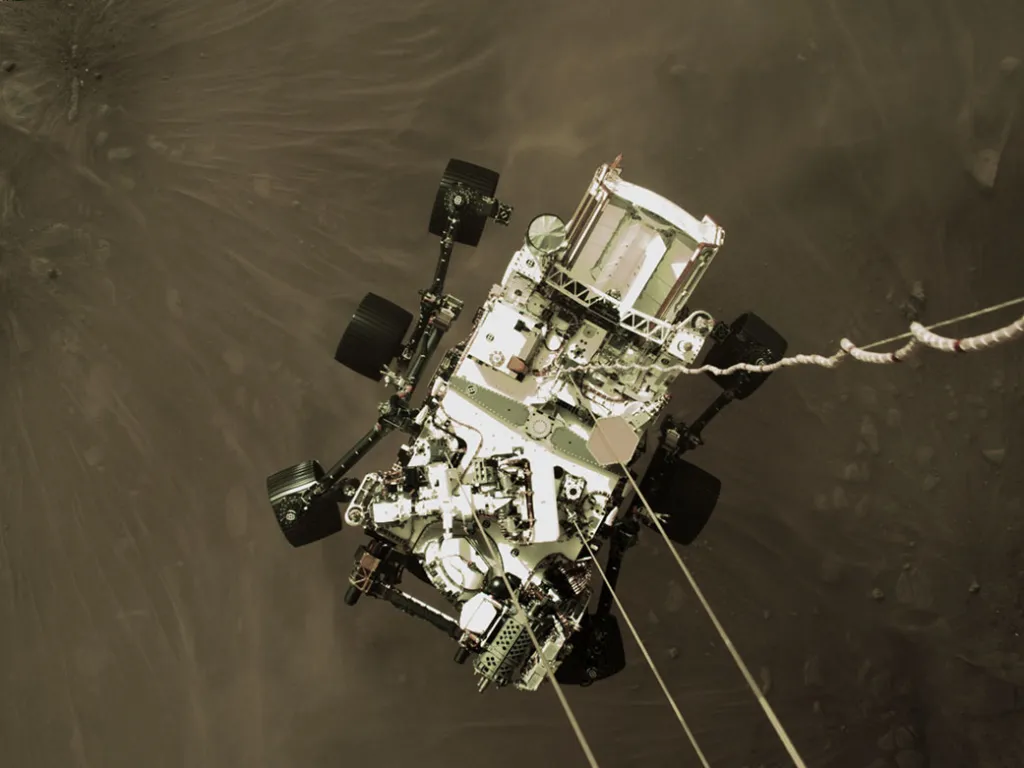 The Mars 2020 descent stage lowers NASA’s Perseverance rover. The six-wheeled rover is seen suspended by cables, with the surface of Mars below.
