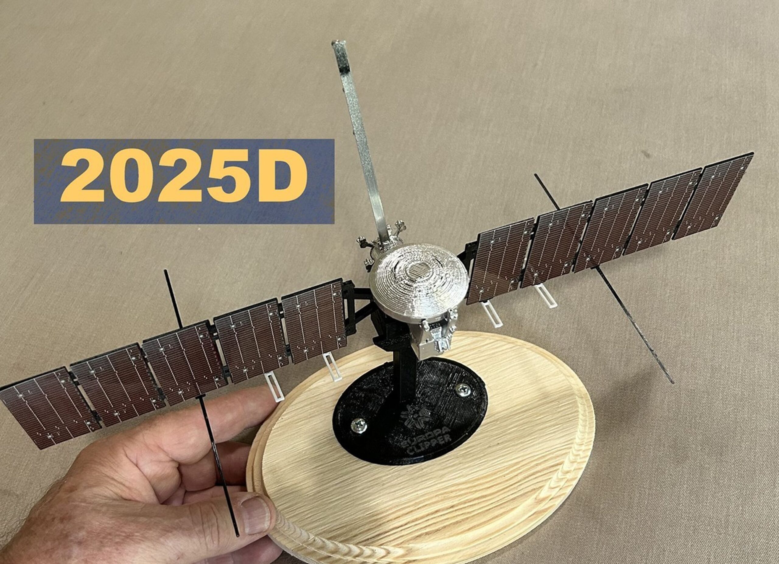 Color photo of a scale model of the Europa Clipper spacecraft. Overlaid on the image is the label "2025D." The model is attached to a small black base, which itself is screwed to an unpainted pine wood base, which is being held by a man's hand coming from the left side of the frame. The model has a small, circular, silver-colored body with two copper-brown solar arrays extending horizontally on either side; each array is about as wide as the silver body, and about four times as long.