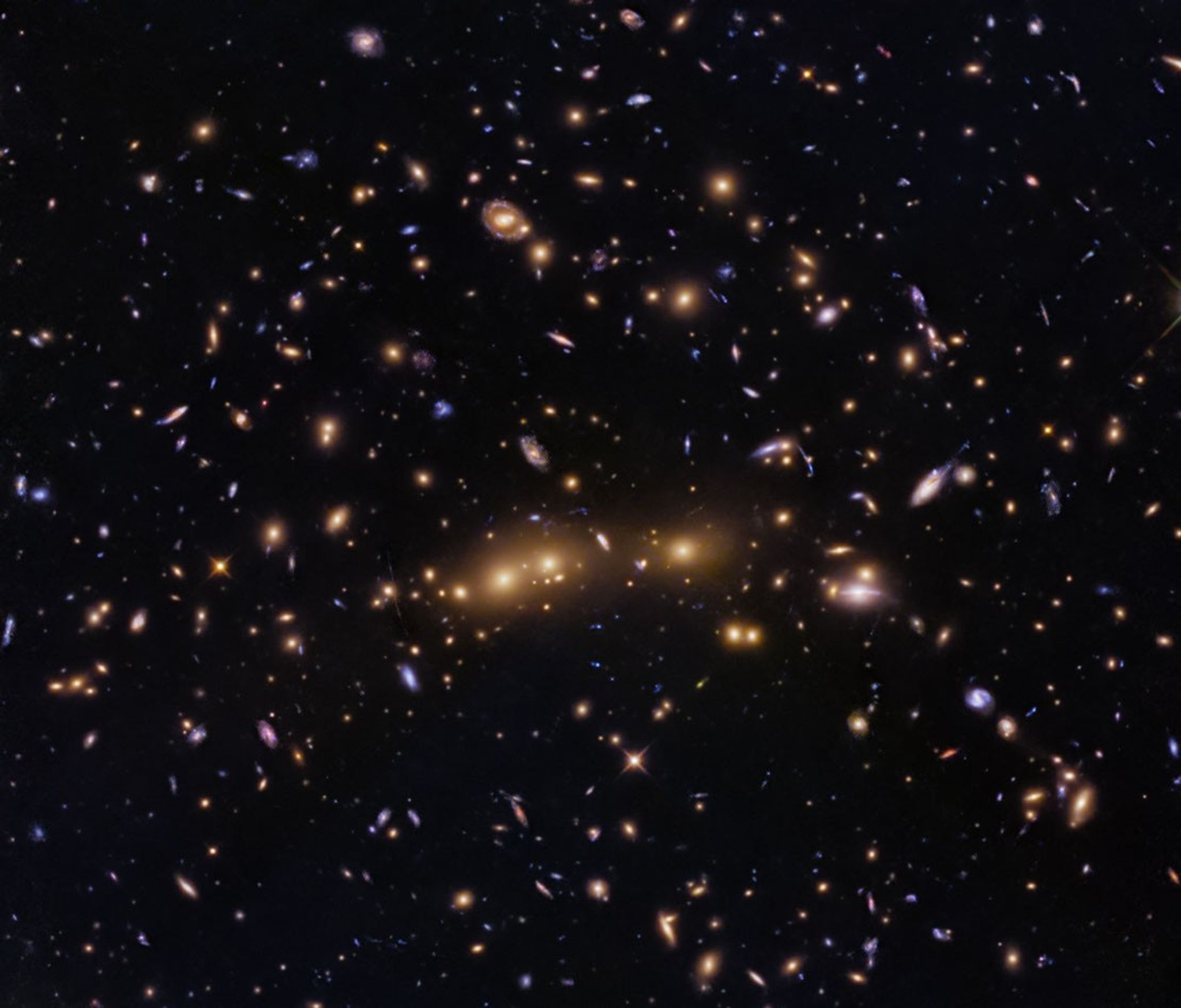 Several massive elliptical galaxies appear to sit in a nearly horizontal line across the center of the image. Numerous spiral, lenticular, and smaller elliptical galaxies dot the space surrounding the giant ellipticals.