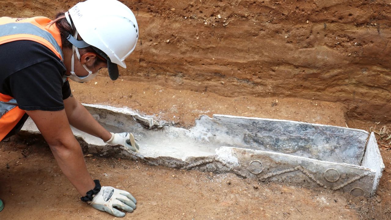 Archaeologist next to the decorated lead coffin partially buried in the ground.