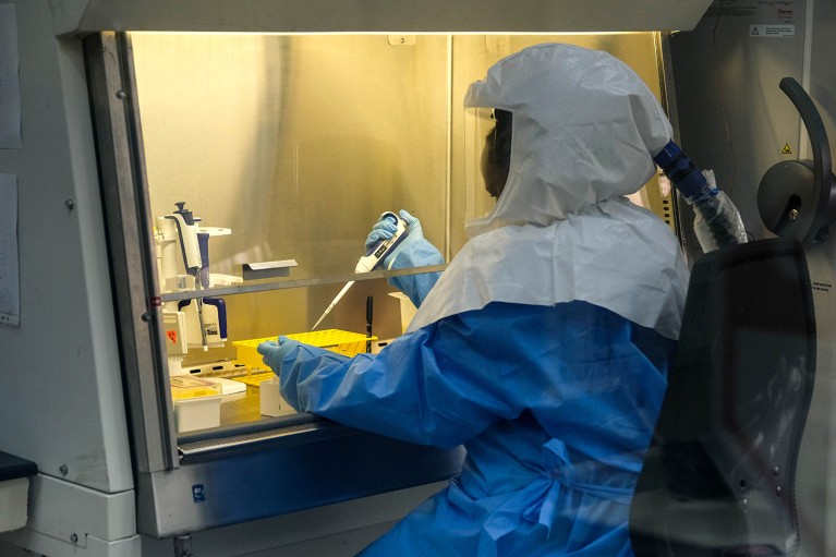 A person in a full biohazard suit pipetting at a biosafety cabinet.