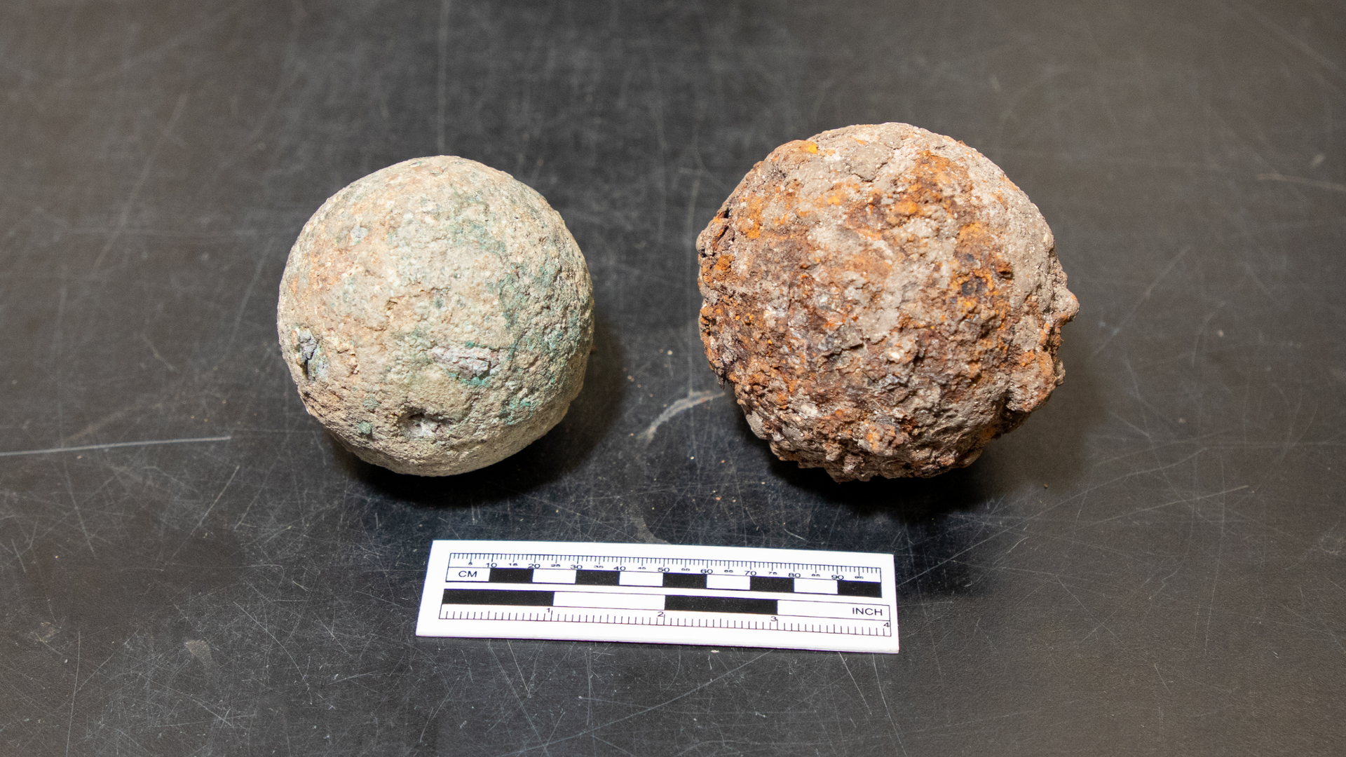 two cannonballs sit side-by-side on a black table with a black-and-white ruler