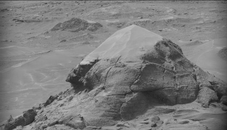 A black-and-white image of a barren Martian landscape featuring a prominent, roughly pyramidal rock outcropping in the foreground. The rock's lower half is rugged with deep cracks and textured layers, while its peak appears lighter and smoother, possibly covered in dust. The surrounding terrain stretches into the background with scattered smaller rocks and another low mound in the distance.