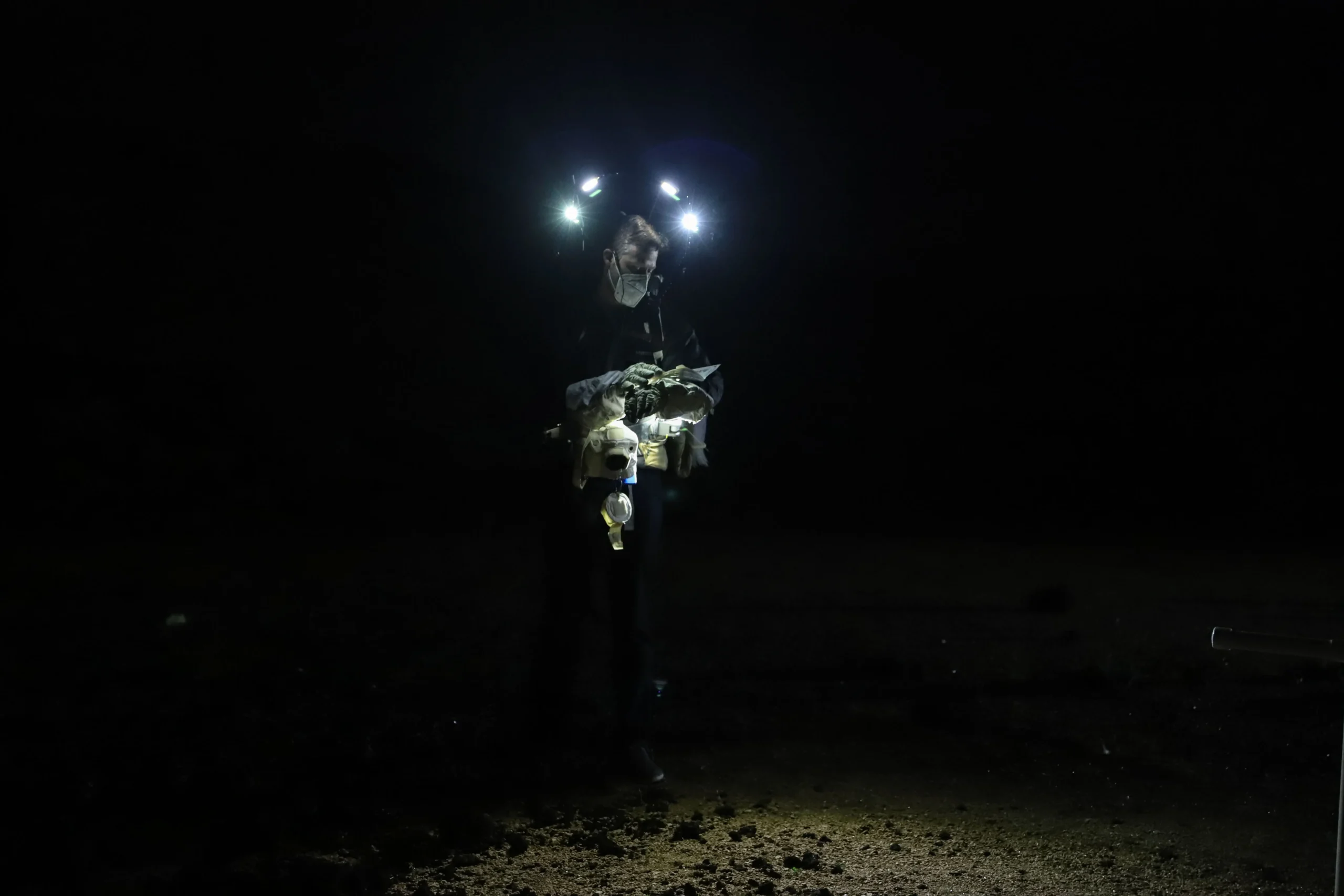 A man wearing an N95 mask, spacesuit gloves, and headlamps studies a booklet of rock samples in the dark.