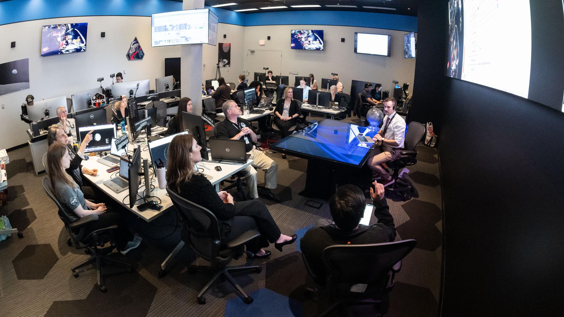 A large operations room filled with about two dozen staff members seated at computer workstations. Rows of desks with multiple monitors face a central area where a person sitting at a glass-topped desk appears to be speaking to the group. Large screens mounted on the walls display space-related imagery and data. Several people are looking towards a large screen hanging on a wall on the right of the image, while others work at their computers. The room is brightly lit, with blue accents along the walls, creating the atmosphere of a space operations or flight control center.