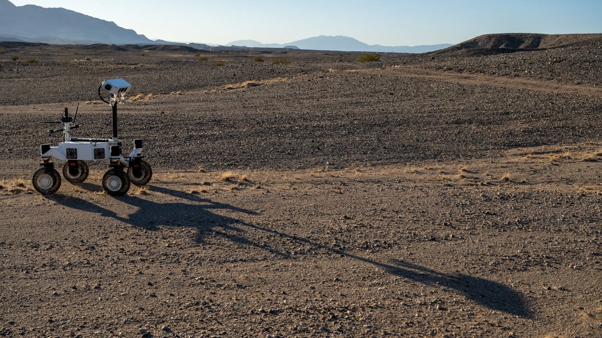 A white four-wheeled rover on the left side of the frame casts a long shadow across barren, brown ground toward the lower right. A few low, scrubby plants dot the desert landscape and mountains rise in the far distance under a pale blue sky.