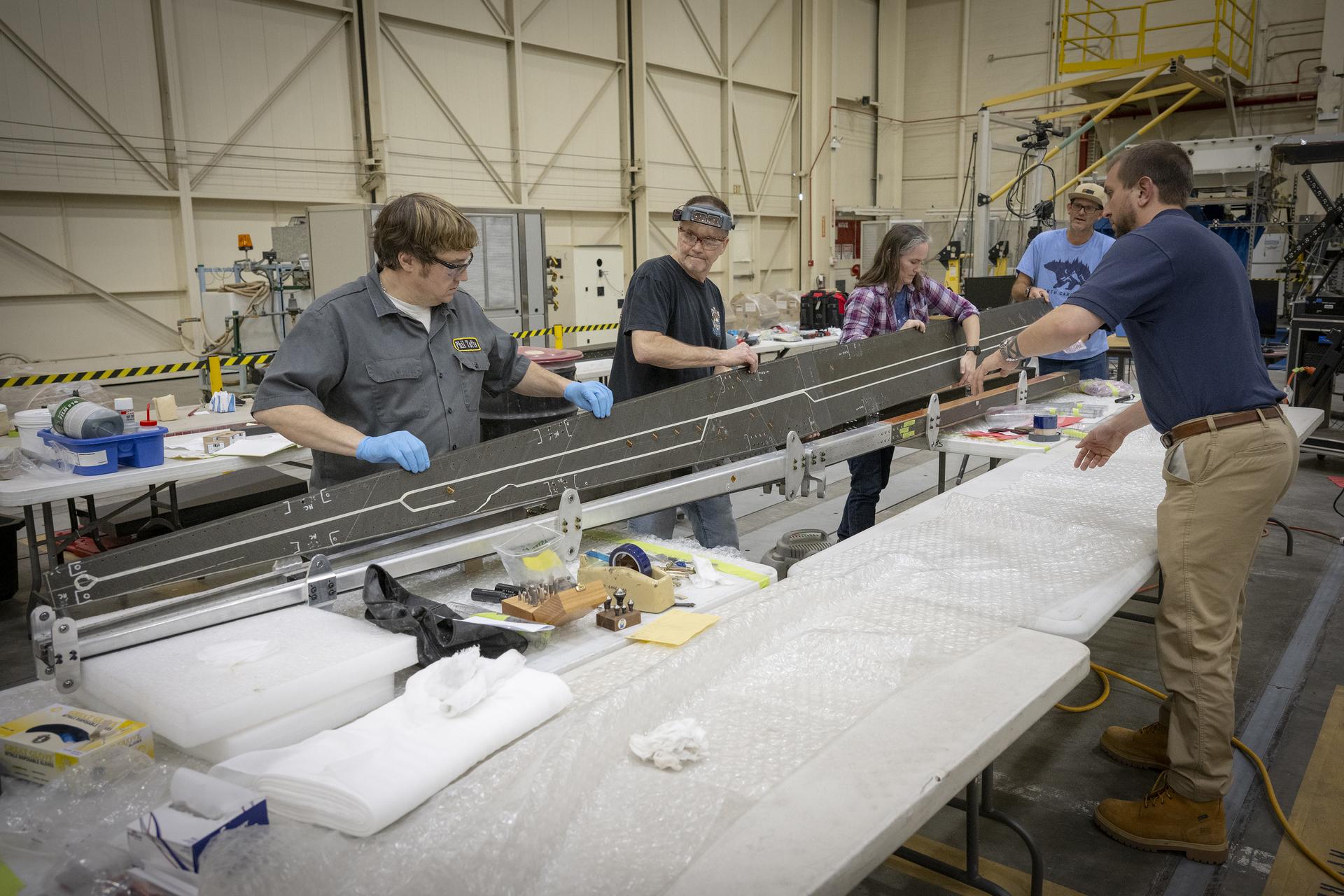 A group of people work together in a large workshop, handling and inspecting a long metallic structure laid across padded tables. Tools, materials, and protective equipment are spread across the workspace.