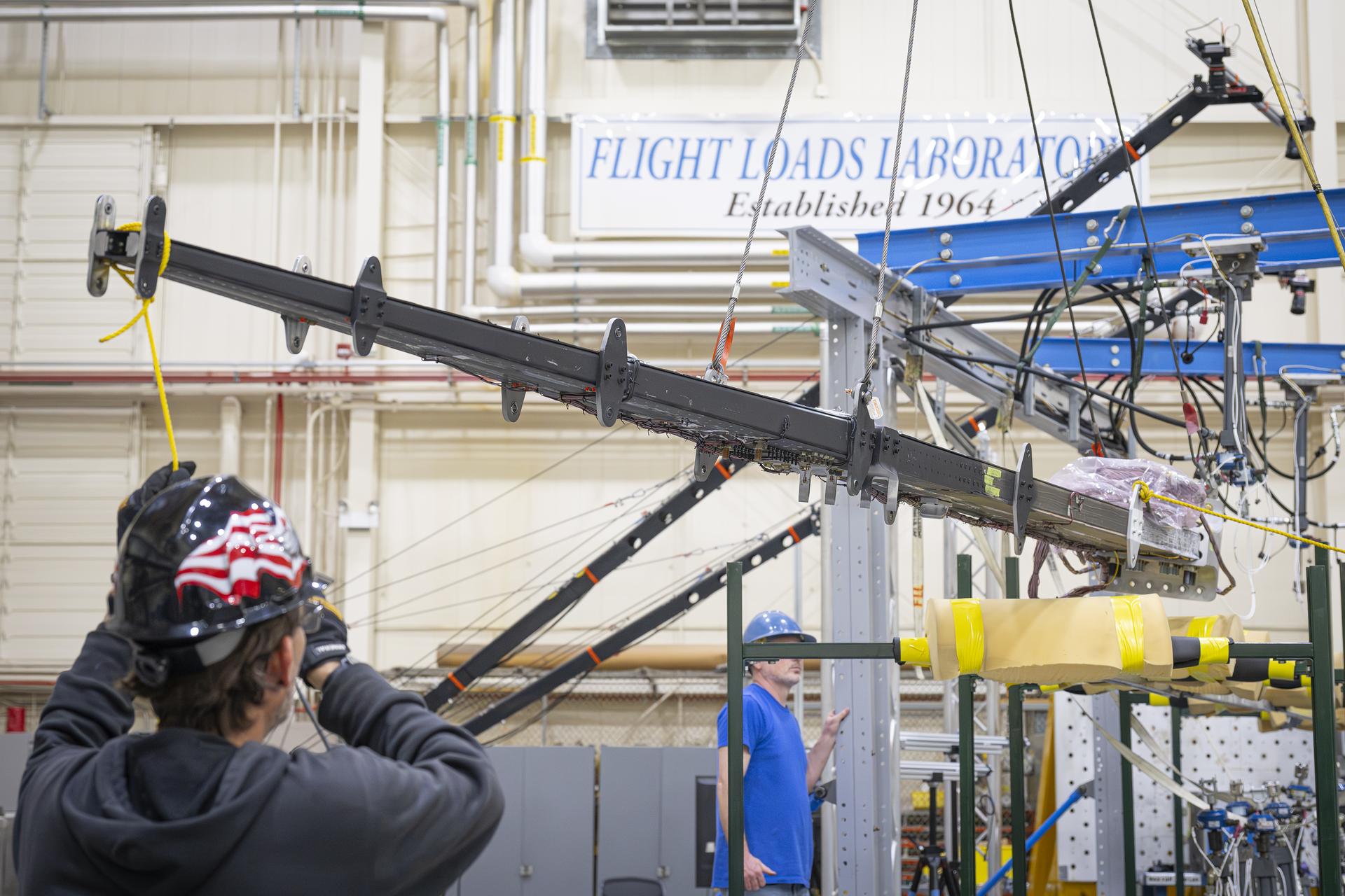 A long beam is suspended in a laboratory while personnel observe and guide its placement. Overhead support equipment, cables, and lab infrastructure surround the test area.
