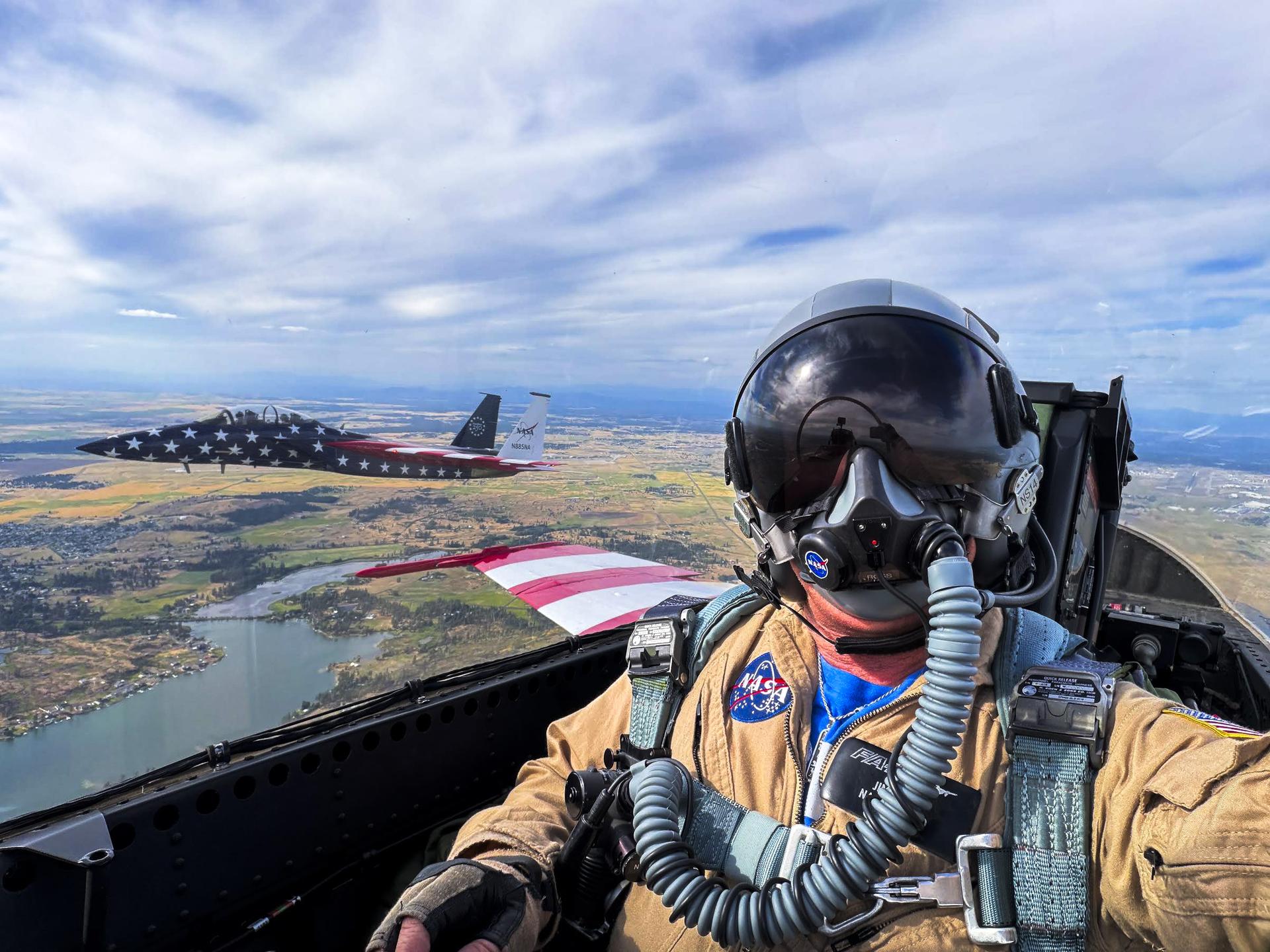 A photographer takes a selfie from the rear seat of a jet aircraft during flight, with another jet visible through the window.