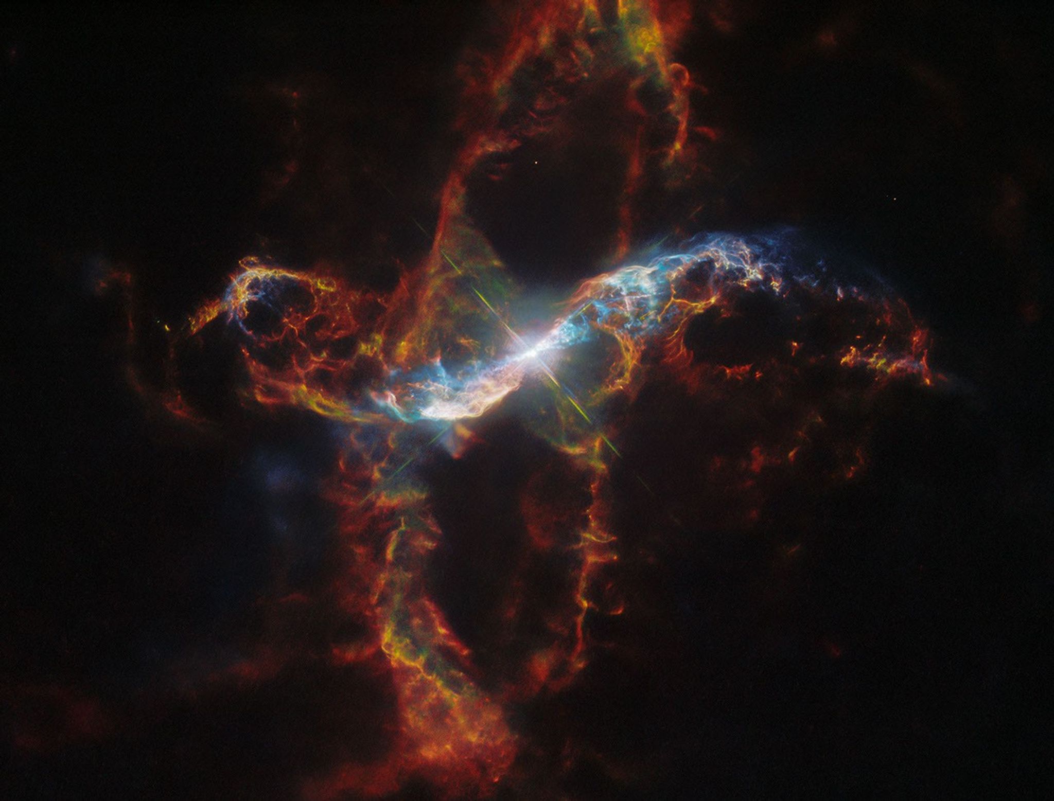 A bright binary star surrounded by a colorful nebula on the black background of space. The star in the center is a large white spot surrounded by a circular glow. It has a large, X-shaped set of diffraction spikes around it. The nebula extends far above, below, left and right of the star in long, arcing shapes made of thin, multicolored filaments — mostly red and greenish colors, but lit in a bright cyan near the star where its light illuminates the gas.