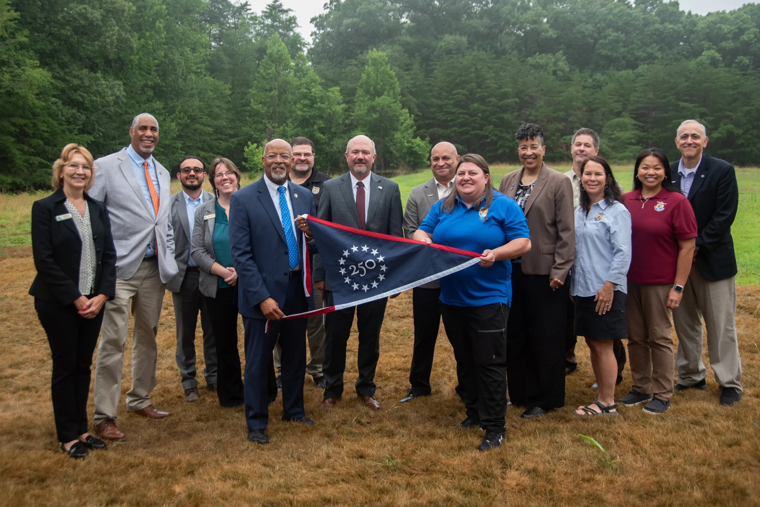a small crowd holds up a triangular pennant flag with "250" on it, with a field and trees in the background
