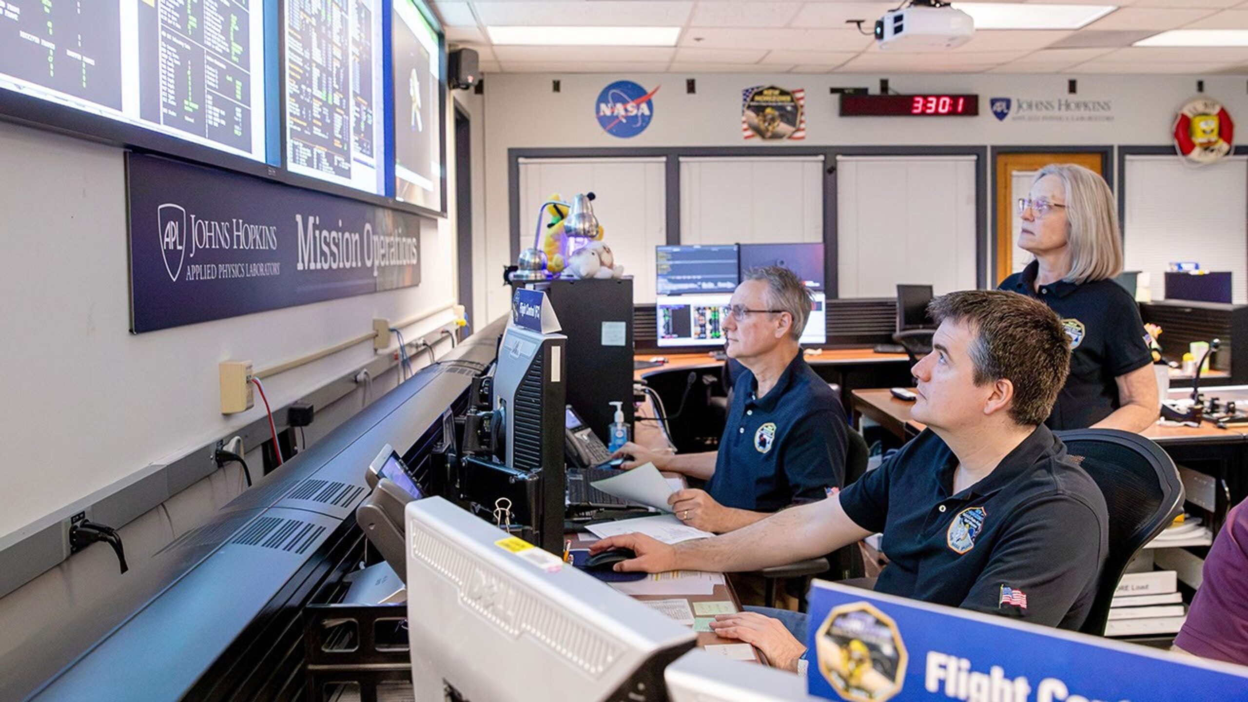 Flight operators at computers in a mission control center monitor spacecraft data on large wall displays.