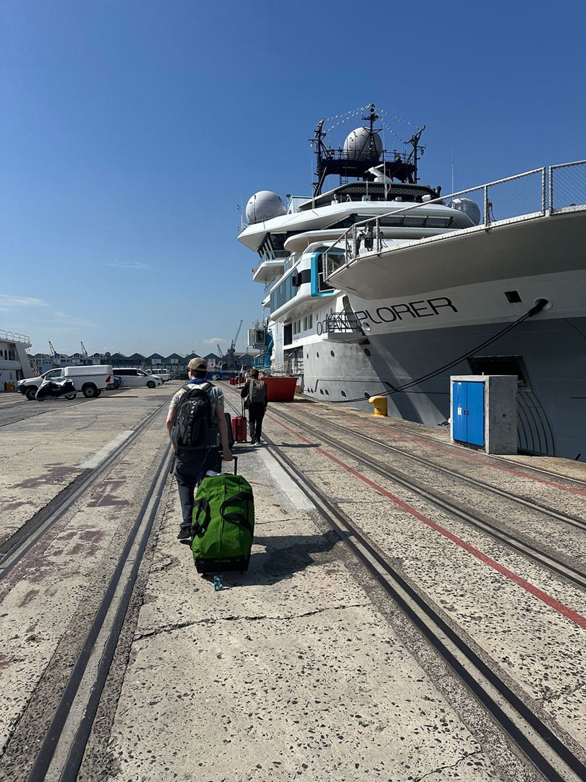 We see two expedition joiners from the rear as they tow their luggage along a concrete wharf lined with tracks for cargo. To the right, the Ocean Explorer ship rises above their heads – a gray-hulled research vessel with a white superstructure, topped by antennas. In the distance stand low buildings and parked vehicles.