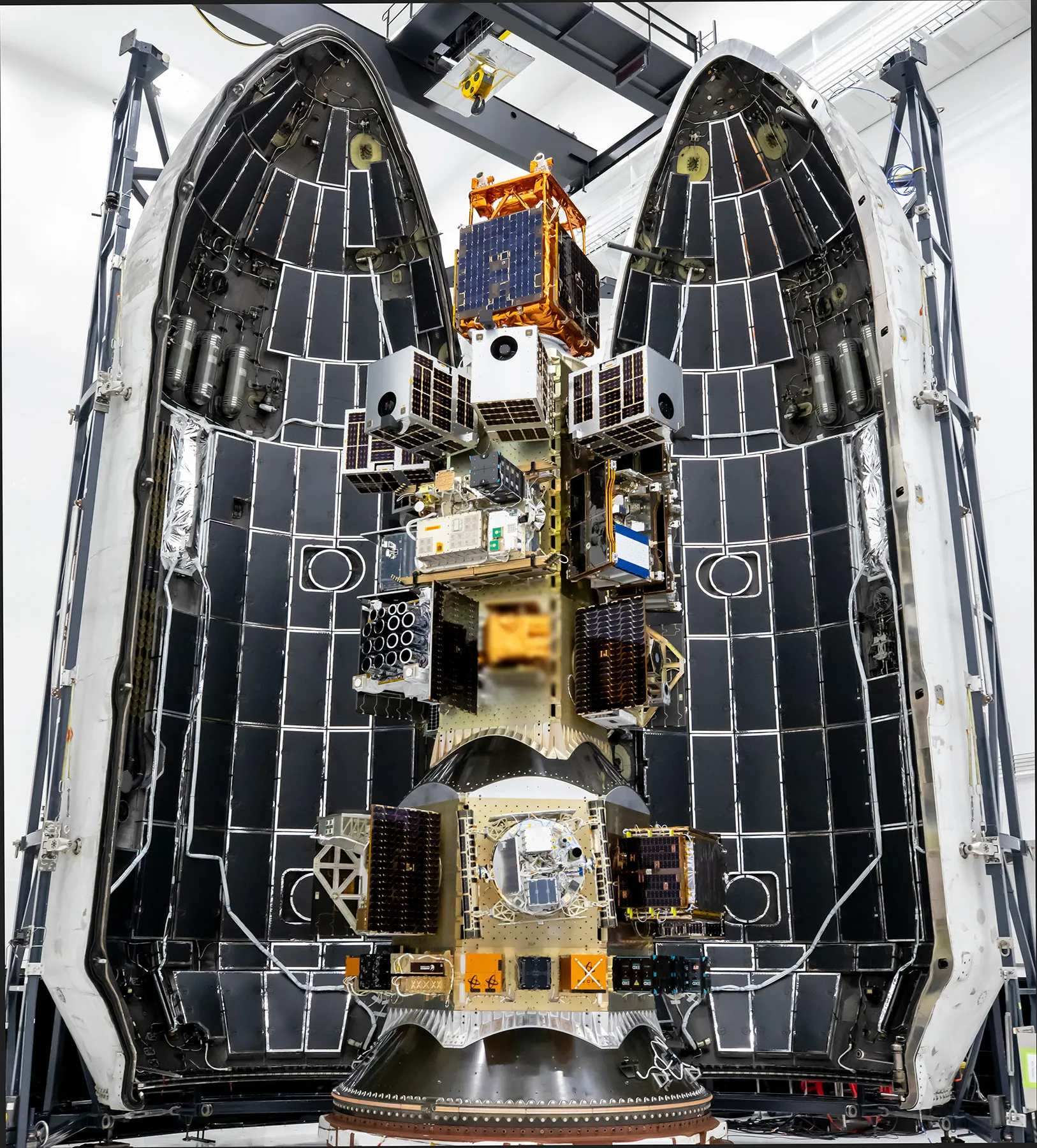 A tall stack of small satellites and payloads is shown mounted on a deployment platform inside an open rocket fairing. The fairing’s two halves are raised, revealing their black internal acoustic panels. The payloads, arranged in multiple tiers, feature boxy shapes with solar panels and wiring. The scene is inside a clean room with support structures surrounding the fairing.