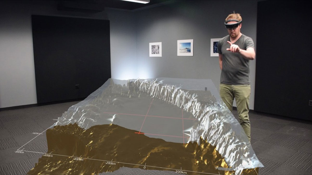 A person stands in an open room wearing virtual reality goggles, with their arm outstretched and index finger and thumb extended. In the foreground, a virtual reality depiction of Antarctic topography and ice are projected atop the photograph.