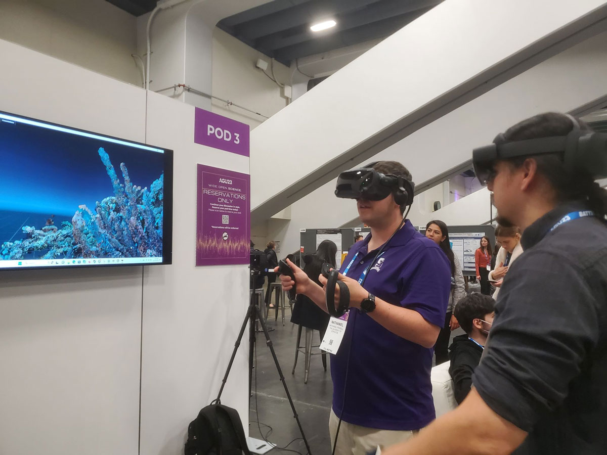 Two people wearing VR headsets stand in front of a television screen showing a VR depiction of a coral reef.