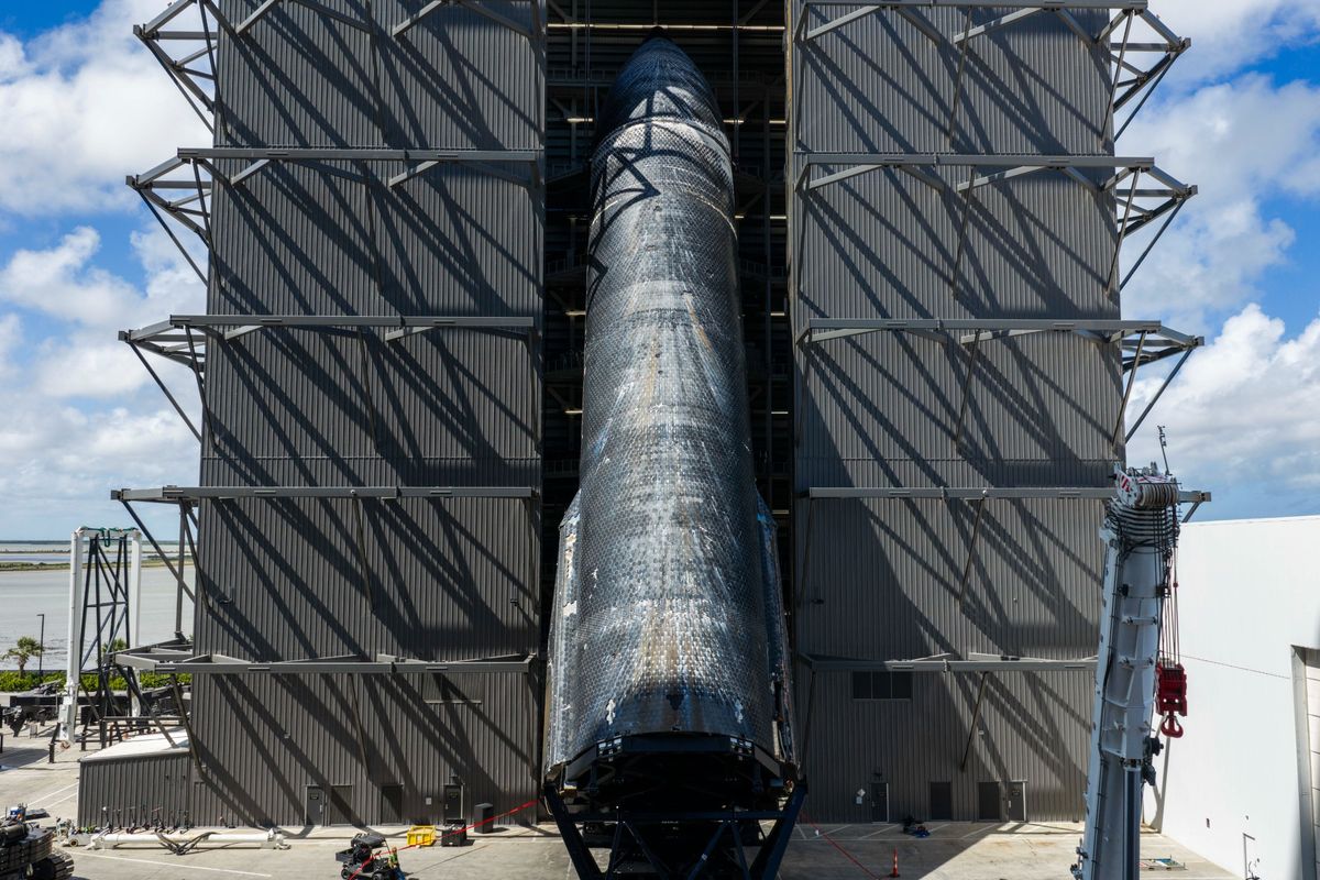 a giant black-tiled rocket is lifted vertical outside a large hanger bay.